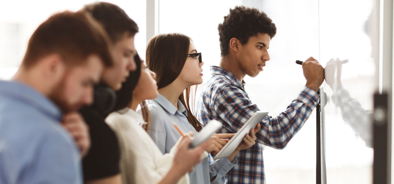 Male high school pupil writing on whiteboard in class, classmates checking task answers